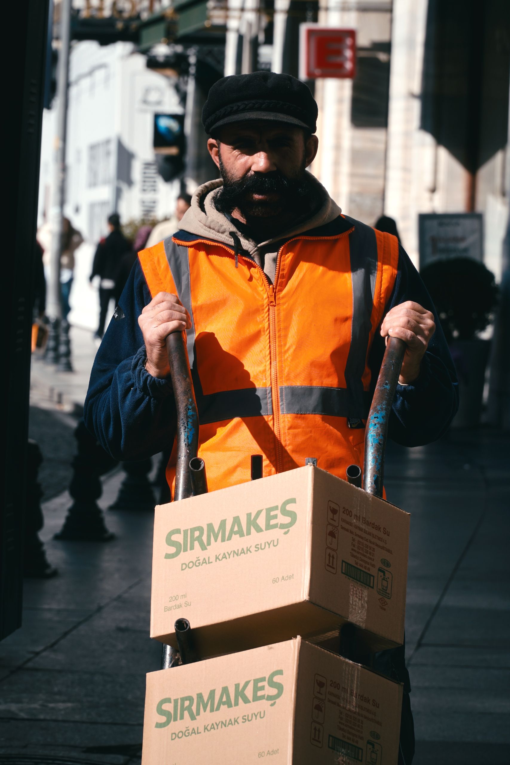 Man wearing an orange vest pushing boxes in a city street, representing urban delivery work.